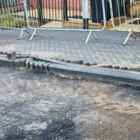A section of newly laid pavement shows damaged kerbing and paving stones.  Loose stones and dirt are scattered nearby. Temporary metal barriers are visible in the background.